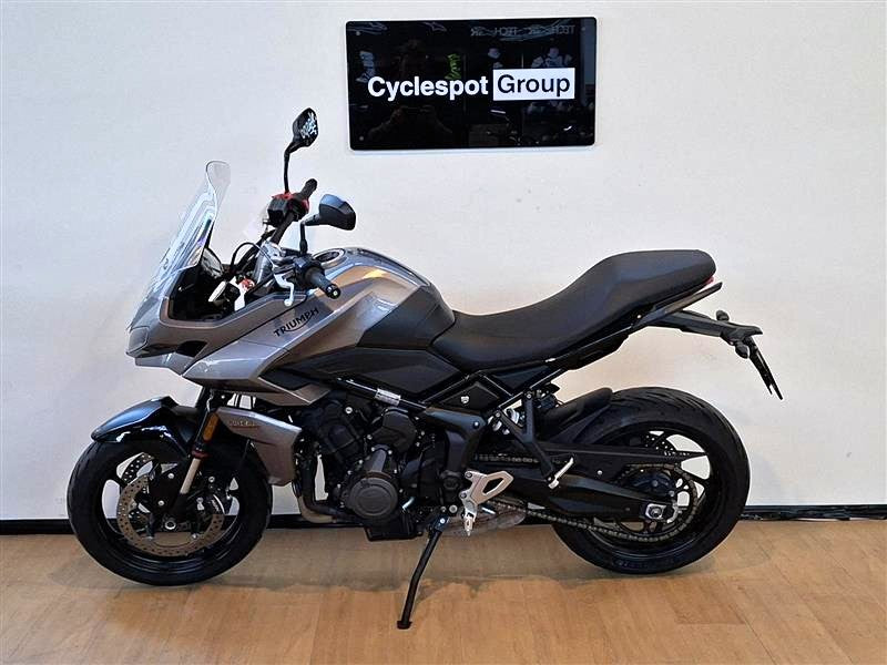 Load image into Gallery viewer, A silver and black 2023 Triumph Tiger Sport 660 Adventure motorcycle is parked indoors on a wooden floor against a white wall, with a 'Cyclespot Group' sign visible on the wall behind it.