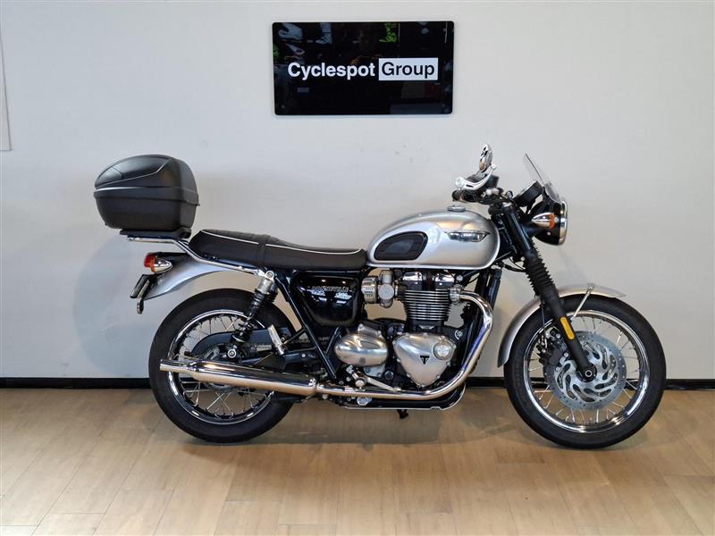 Load image into Gallery viewer, A silver and black 2018 Triumph Bonneville T100 motorcycle with a top case, displayed in profile against a light gray wall with a 'Cyclespot Group' sign above it.