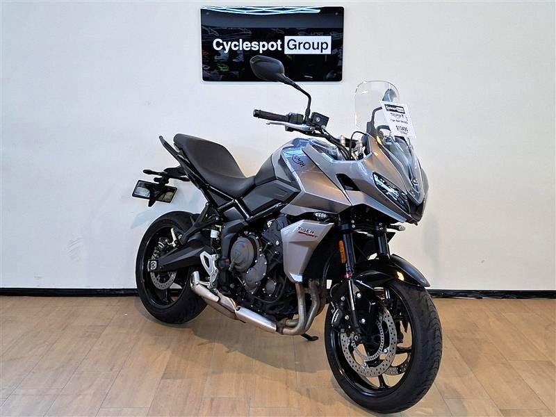 Load image into Gallery viewer, A silver and black 2023 Triumph Tiger Sport 660 motorcycle is parked indoors on a light wood floor against a white wall, with a 'Cyclespot Group' sign visible above it.