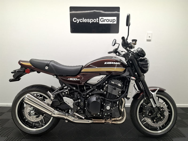Load image into Gallery viewer, A Kawasaki Z900RS motorcycle in a dark maroon and black color scheme with gold stripes, displayed indoors against a white wall with a 'Cyclespot Group' sign above it.
