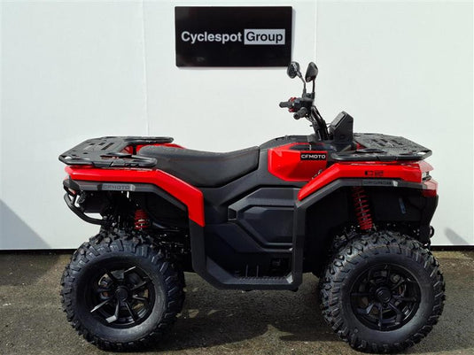 A red and black 2026 CFMOTO CFORCE C5 EPS with large knobby tires, rear cargo racks, and a black seat, photographed in profile against a white wall with a 'Cyclespot Group' sign above it.