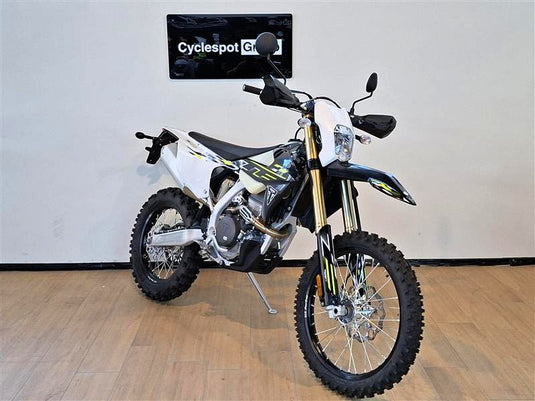 A white and black Triumph TF 250 E dirt bike with yellow accents, displayed on a stand in an indoor showroom with a wood floor and a wall sign reading 'Cyclespot Gr'.