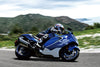 A blue and white 2026 Suzuki Hayabusa GSX 1300 RR Special Edition supersport motorcycle with a rider in full racing gear, captured in motion on a paved road with green hills and a cloudy sky in the background.