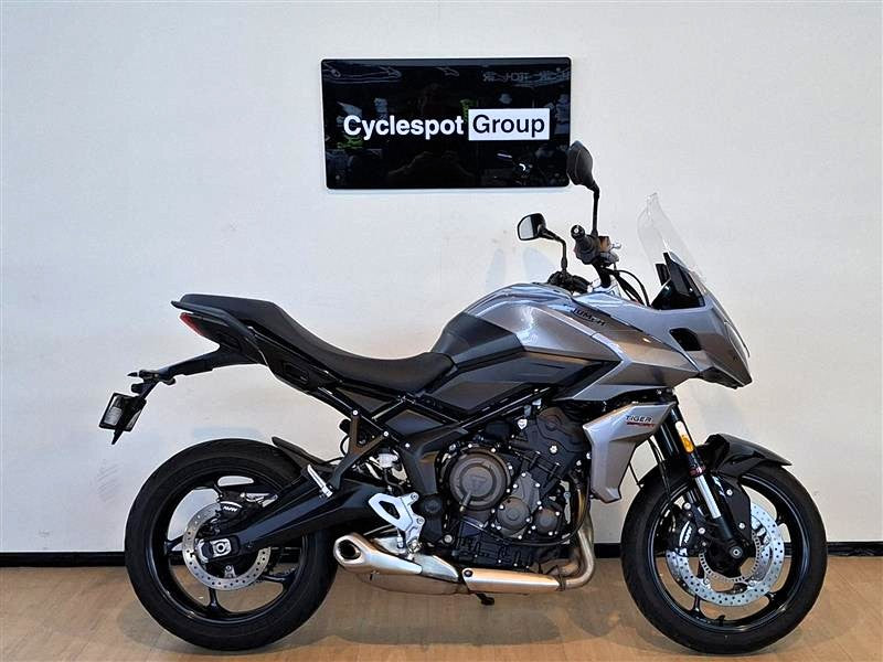 Load image into Gallery viewer, A silver and black  2023 Triumph Tiger Sport 660 motorcycle is parked indoors against a white wall with a 'Cyclespot Group' sign above it. The bike is shown in full side profile with its front wheel turned slightly inward.
