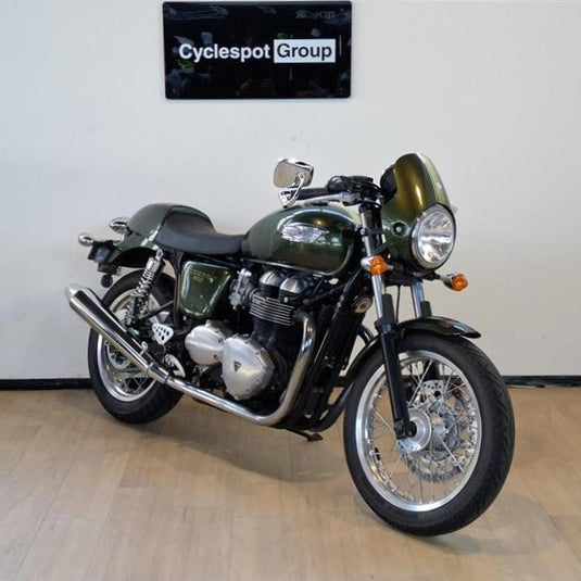 A classic-style 2013 Triumph Thruxton 900 motorcycle with a dark green metallic finish and chrome engine components, parked indoors on a light wood floor against a white wall with a 'Cyclespot Group' sign above it.