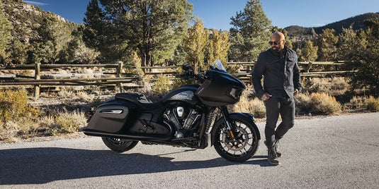 A man in black riding apparel stands beside a black 2026 Indian Challenger Dark Horse motorcycle on an outdoor paved road with trees and hills in the background.