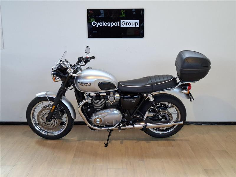 Load image into Gallery viewer, A silver 2018 Triumph Bonneville T100 motorcycle with a black seat and black top case, parked indoors on a light wood floor against a white wall with a 'Cyclespot Group' sign above.
