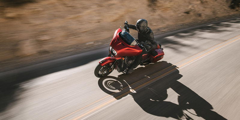 Load image into Gallery viewer, A red 2026 Indian Chieftain Dark Horse motorcycle with a rider in black gear traveling on a paved road with dry brush in the background, captured from a dynamic side angle with motion blur.
