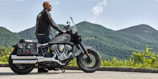 A man wearing a black leather jacket and jeans sits on a black and silver 2026 Indian Super Chief Dark Horse motorcycle with saddlebags, parked outdoors on a paved road with green mountains in the background.