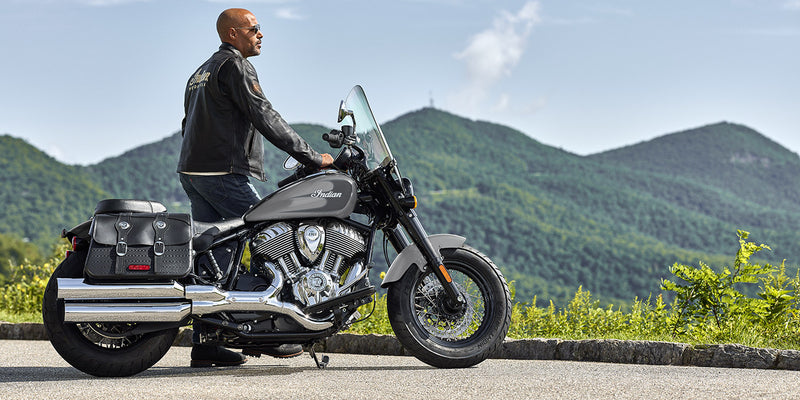 Load image into Gallery viewer, A man wearing a black leather jacket and jeans sits on a black and silver Indian Super Chief motorcycle with saddlebags, parked outdoors on a paved road with green mountains in the background.
