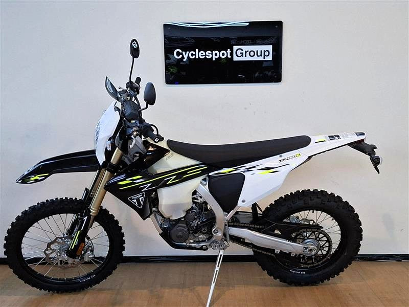 Load image into Gallery viewer, A white and black Triumph TF 450-E dirt bike with yellow accents, displayed on a kickstand in an indoor showroom setting with a wall sign reading 'Cyclespot Group'.
