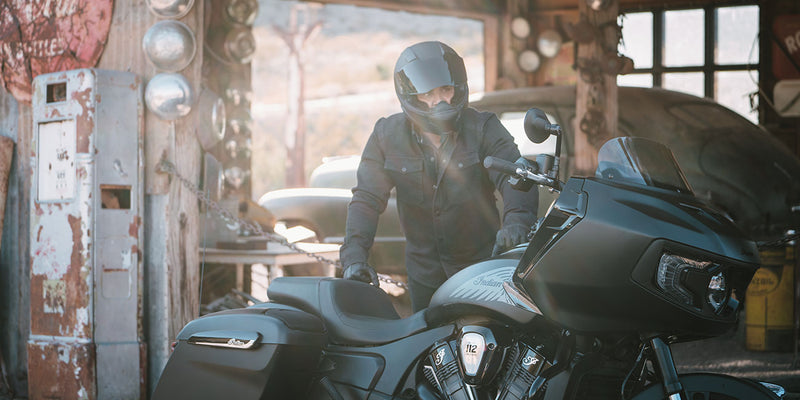 Load image into Gallery viewer, A person in a black helmet and dark jacket stands beside a large black 2026 Indian Challenger Limited in a rustic, sunlit garage or workshop with vintage equipment and wooden posts.
