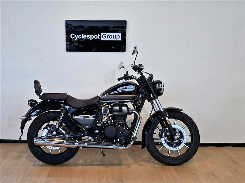 Load image into Gallery viewer, A black and silver Royal Enfield cruiser motorcycle with a brown seat, displayed in profile on a wooden floor against a light gray wall. A 'Cyclespot Group' sign is mounted on the wall above the motorcycle.
