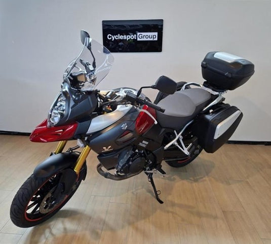 A Suzuki DL1000 V-Strom adventure-touring motorcycle with a tall windscreen, saddlebags, and a top box, parked indoors on a wooden floor in front of a white wall with a Cyclespot Group sign.