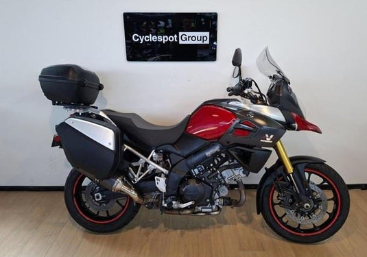 A red, black, and silver Suzuki DL1000 V-Strom Adventure Tour Sport motorcycle with black panniers and a top case, parked indoors against a white wall with a 'Cyclespot Group' sign above it.