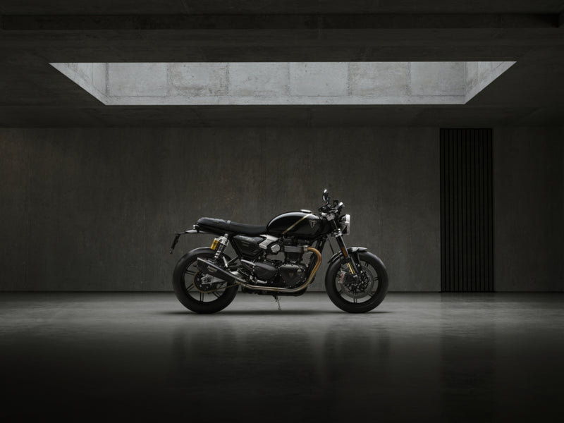 Load image into Gallery viewer, A black and silver 2027 Triumph Speed Twin 1200 TFC motorcycle is parked in profile against a dark concrete wall in a minimalist, moody studio setting with a rectangular skylight overhead.
