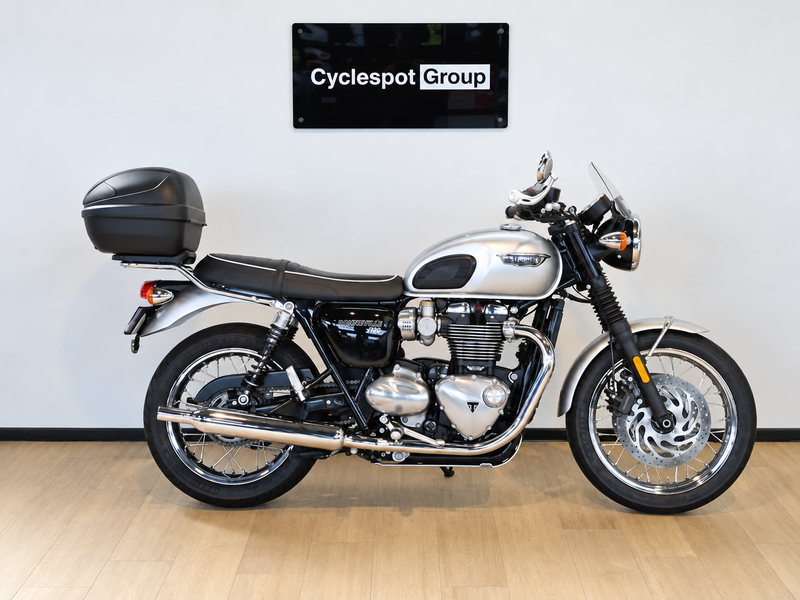 Load image into Gallery viewer, A silver and black Triumph Bonneville T100 motorcycle with a black top case, displayed in profile on a light wood floor against a white wall with a 'Cyclespot Group' sign above it.

