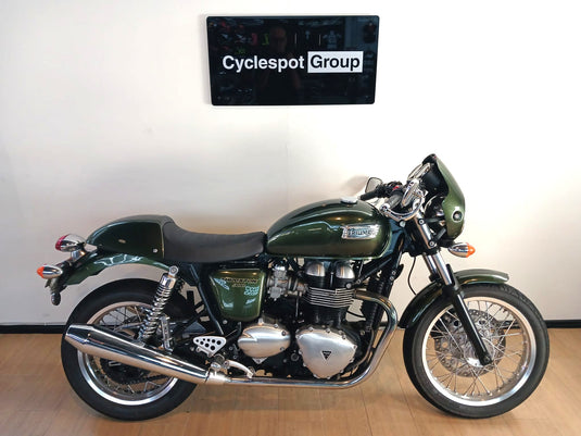 A dark green 2013 Triumph Thruxton 900 motorcycle with chrome details, parked indoors against a white wall with a 'Cyclespot Group' sign above it.