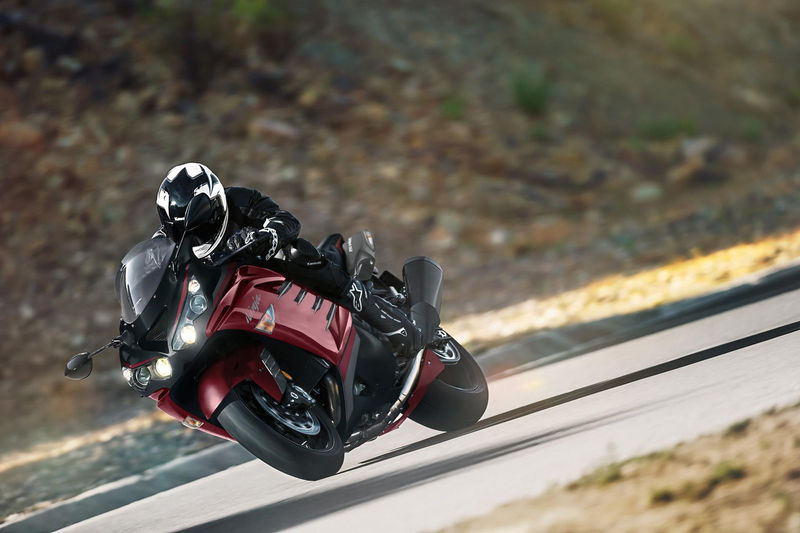 Load image into Gallery viewer, A red and black 2025 Kawasaki Ninja ZX 14 R SE motorcycle with a rider in black gear and a white helmet, leaning sharply during a turn on a paved road with a blurred natural background.
