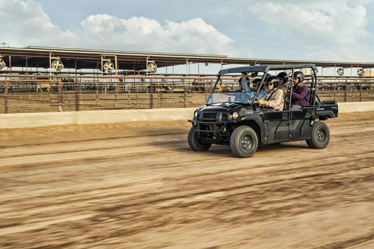 A black four-seater utility off-road vehicle with passengers driving on a dirt track, with a large covered structure in the background under a cloudy sky.