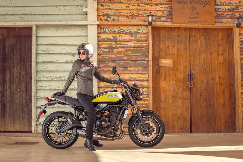 Load image into Gallery viewer, A person wearing a black leather jacket, black pants, and a white helmet sits on a yellow and black Kawasaki Z650RS motorcycle in front of a rustic wooden building.
