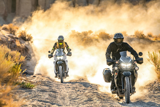 Two motorcyclists riding off-road 2026 Kawasaki KLR650 Adventure (LAMS) adventure bikes on a dusty dirt trail in a desert landscape, with one rider in the foreground and another following behind, kicking up a large cloud of golden dust.
