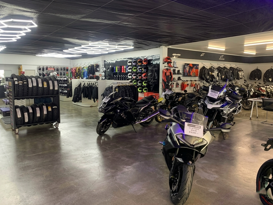 Inside Cyclespot Taranaki, featuring a wide selection of motorcycles for sale and the largest motorcycle accessories department in the region. The modern showroom includes a tyre rack, helmet wall, and riding gear under distinctive geometric lighting.