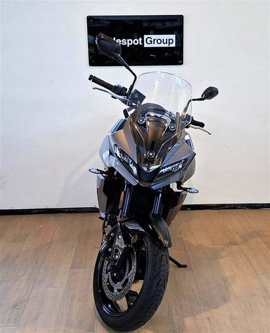 A grey  2023 Triumph Tiger Sport 660  motorcycle is parked indoors on a wooden floor against a white wall, with a 'spot Group' sign visible above it.