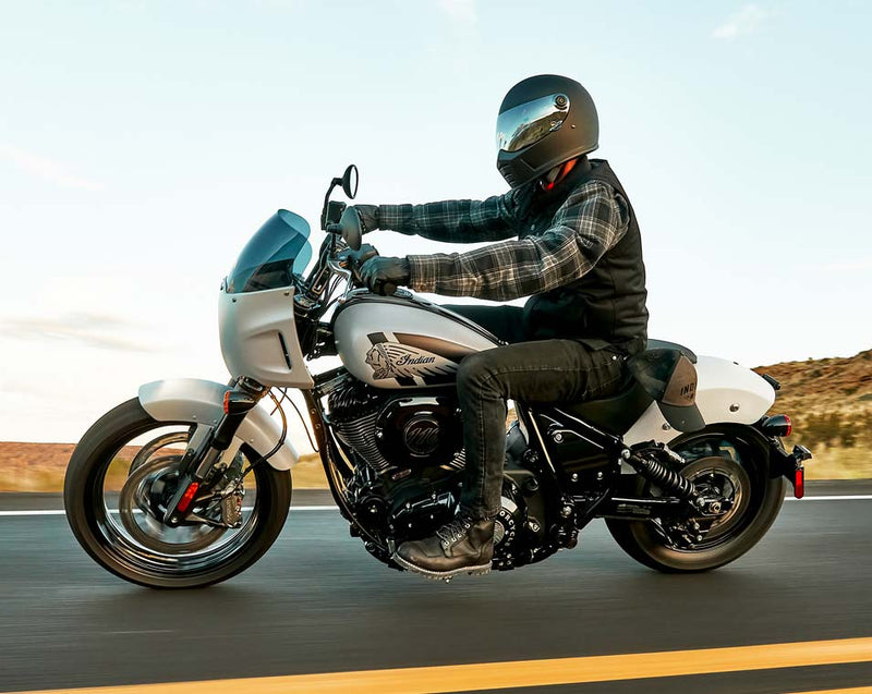 Load image into Gallery viewer, A person wearing a black helmet, plaid shirt, and black vest rides a white and black Indian Sport Chief cruiser motorcycle on a paved road with a desert landscape in the background.

