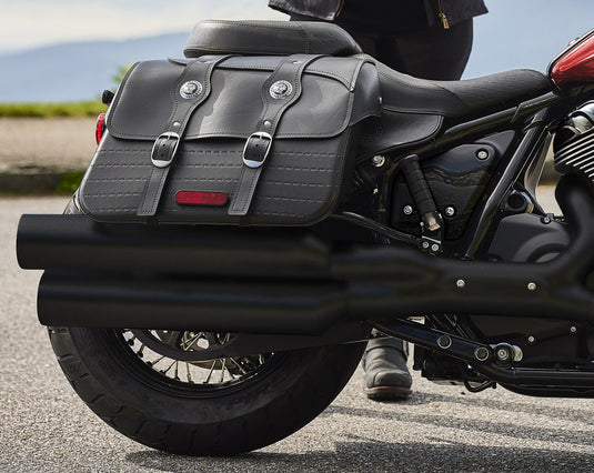A close-up side view of a black leather motorcycle saddlebag bag mounted on the rear of an Indian Super Chief cruiser motorcycle, with a person's leg visible standing beside it on an outdoor paved surface.