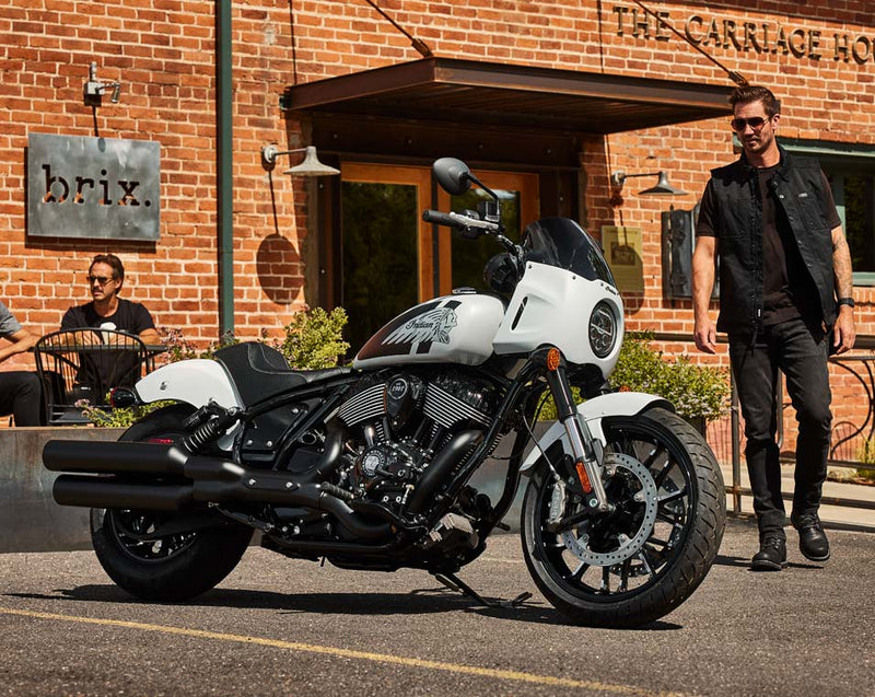 Load image into Gallery viewer, A white and black Indian Sport Chief cruiser motorcycle is parked outdoors in front of a brick building with a man walking beside it and another seated person in the background. The motorcycle has a white fuel tank and body panels with black engine, exhausts, seat, and wheels.
