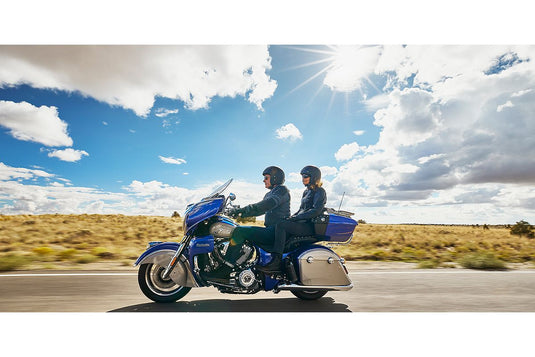 A blue and silver Roadmaster touring motorcycle with two riders traveling on a paved road under a bright blue sky with sun rays and clouds.