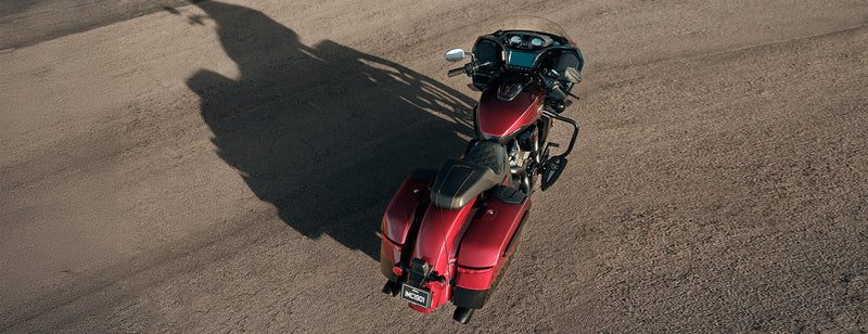 Load image into Gallery viewer, A red 2026 Indian Challenger 125th Anniversary motorcycle with black saddlebags and a windshield, photographed from a high-angle aerial perspective on a dusty, light-brown gravel surface.
