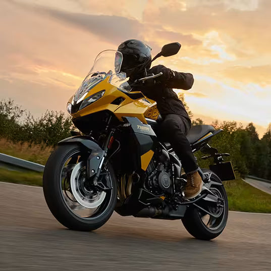 Load image into Gallery viewer, A person wearing a black helmet and riding gear sits on a yellow and black Triumph Tiger 800 motorcycle, captured from a low angle on a road during a warm sunset sky.
